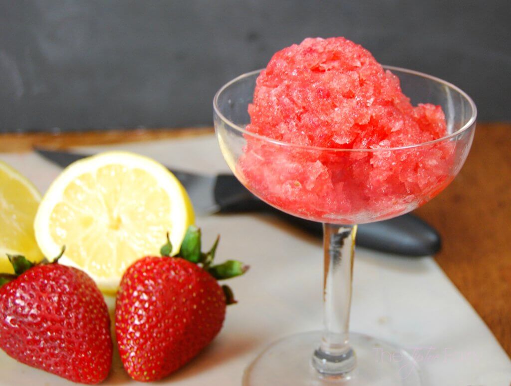 A scoop of strawberry lemonade granita in a champagne glass with a sliced lemon and strawberries on a cutting mat