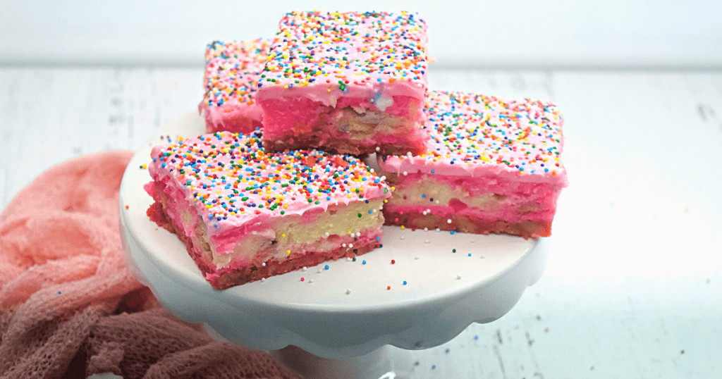 Cut squares of animal cracker cheesecake on a whtie cake plate where you can see layers of swirled pink and white cheesecake. 