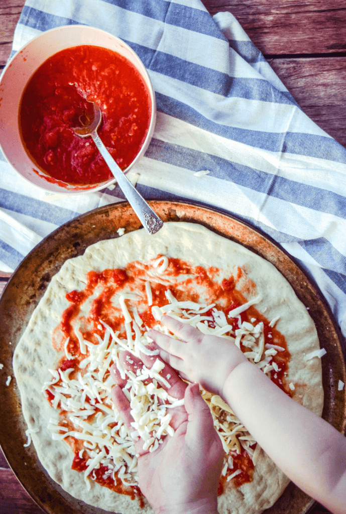 Bowl of pizza sauce and pizza being made with sauce and mozzarella cheese with a mommy hand a kid hand sprinkling the cheese. 