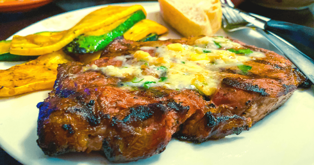 A juicy steak with melted steakhouse butter over the top with a piece of bread and strips of grilled zucchini in the background.