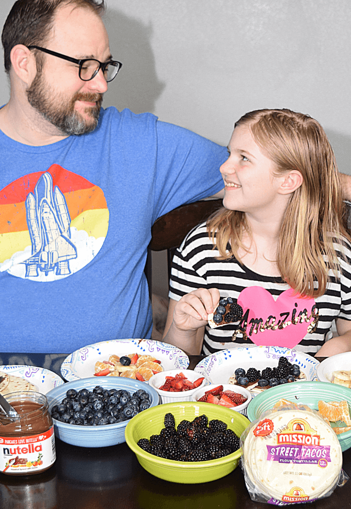 Daddy and daughter smiling at one another as they make funny face fruit pizzas. 