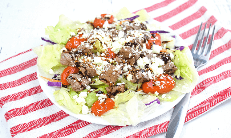 A steak salad with lettuce, steak, roasted grape tomatoes, topped in feta cheese and balsamic glaze. 