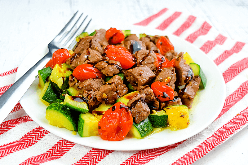 A close up image of the finished meal with a white plate full of cooked zucchini, sirloin steak, and roasted tomatoes drizzled in garlic butter and balsamic glaze. 