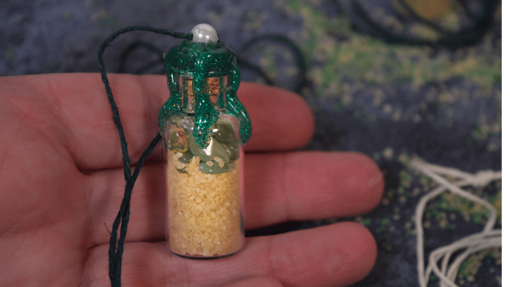 A hand holding a finished spell bottle necklace with a pearl in the top of the hot glue and a green cord threaded through the pearl to make the diy halloween spell bottle necklace
