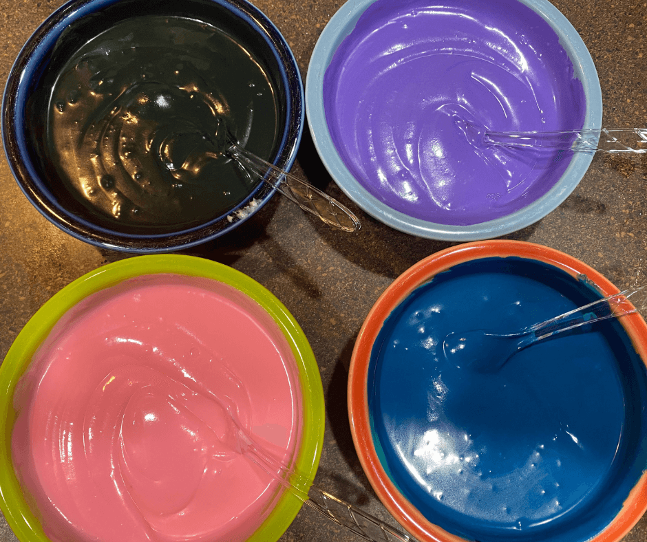 Four bowls of cheesecake batter with gel food coloring in black, dark blue, pink, and purple