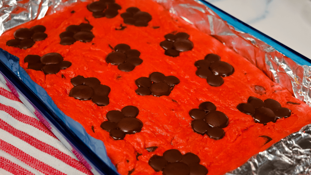 A blue casserole dish lined in foil with finished baking red velvet blondie bars with chocolate pawprints on top. 