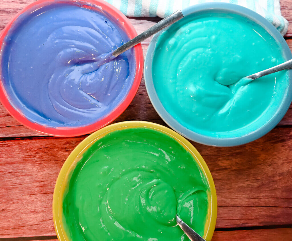 Three bowls with purple, blue, and green food coloring cheesecake batter.