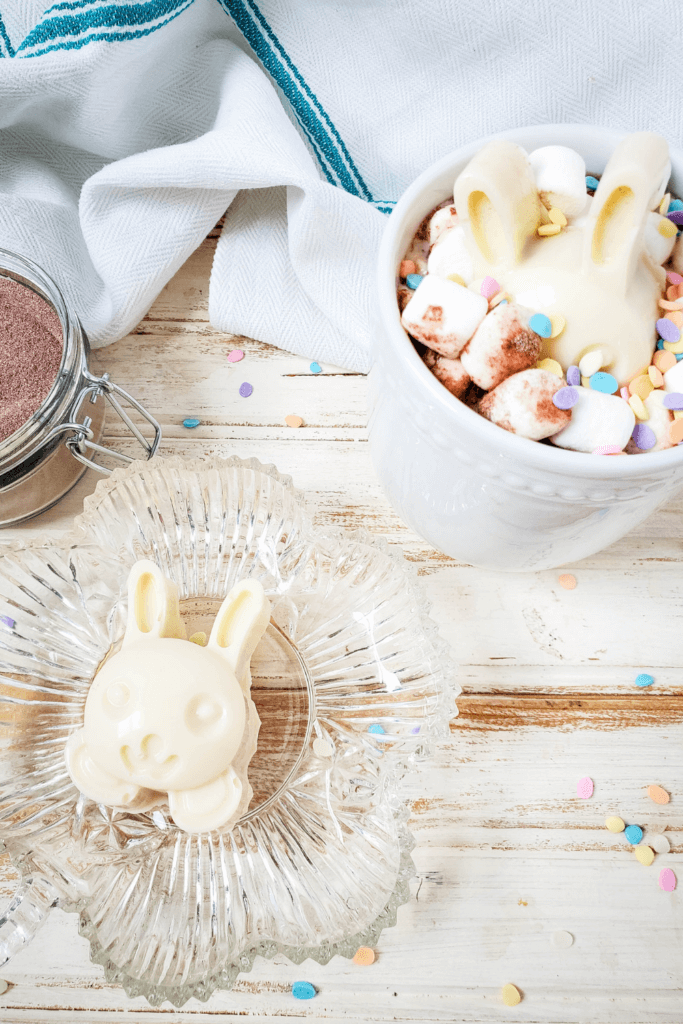 A finished easter bunny hot cocoa bomb on a plate with a mug of hot cocoa with marshmallows, sprinkles, and a hot cocoa bomb. 