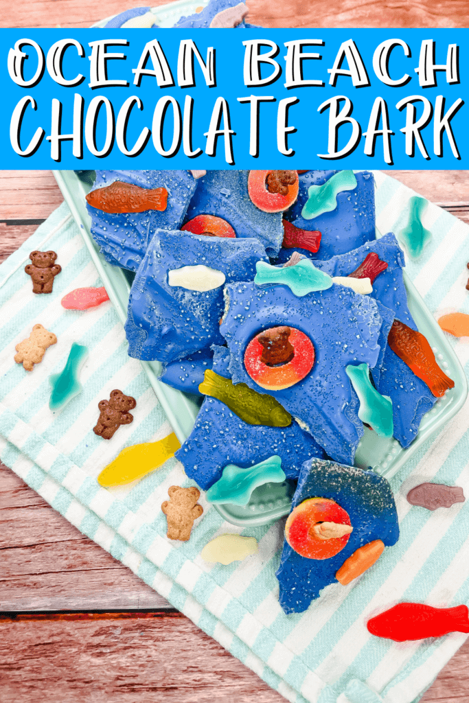 Flat lay of the ocean beach chocolate bark with the pieces on a light blue platter on a striped blue towel surrounded by teddy grahams and gummy fish. 