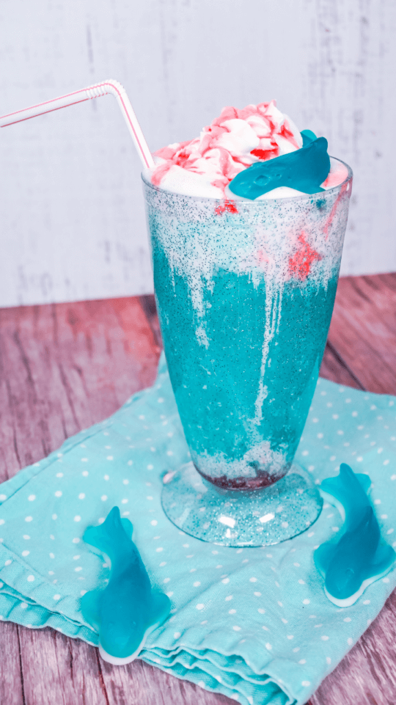 Shark Attack Ice Cream Float with red strawberry syrup and jumbo gummy sharks on a blue polka dot napkin