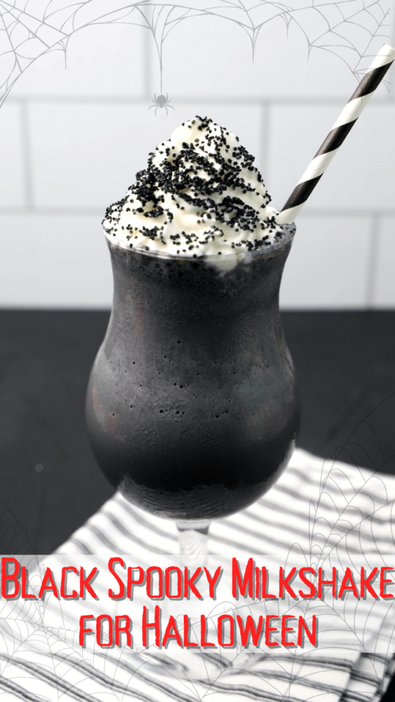 A Black Spooky Milkshake for Halloween topped with whipped cream and black sprinkles on a black striped towel surrounded by spiderwebs.