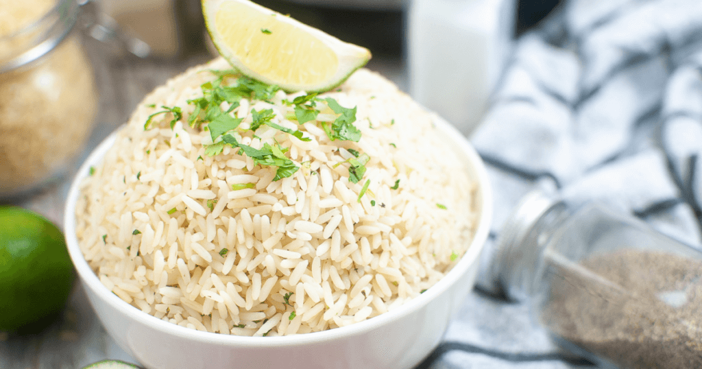 Close up image of Cilantro Lime Rice with a lime wedge on top of it in a white bowl surrounded by spices and a black and white striped towel