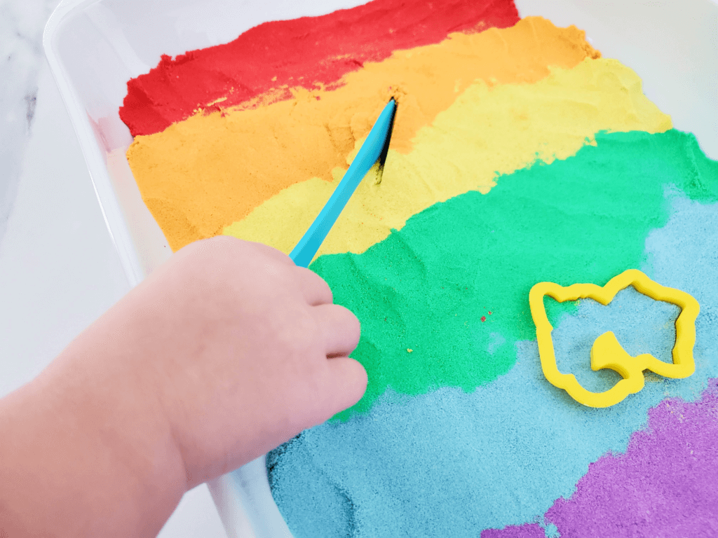 A child's hand holding a plastic knife cutting into the rainbow kinetic sand. 