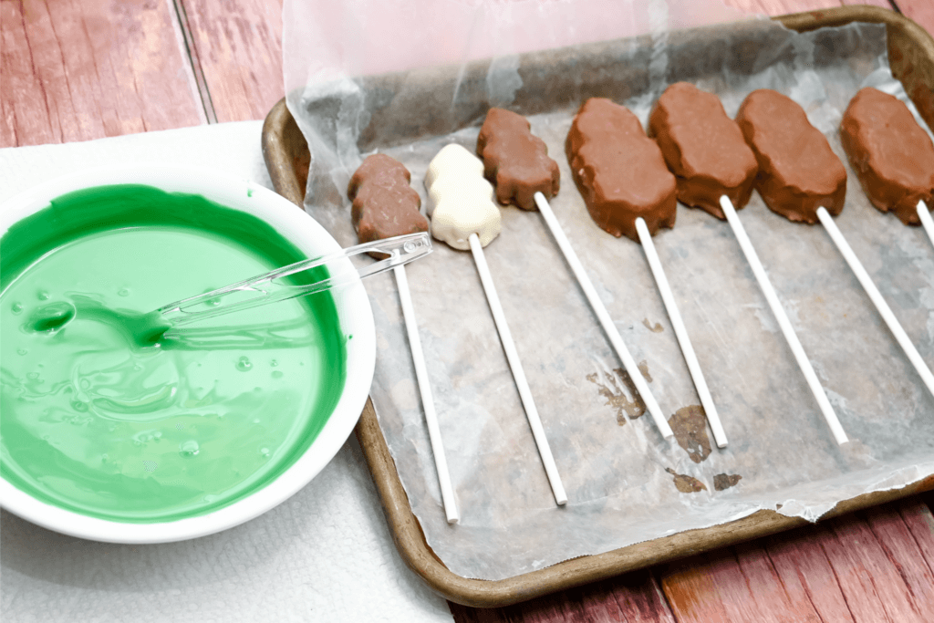 The green candy melts now melted in a bowl with a spoon next to the Reese's Trees on a cookie sheet with wax paper