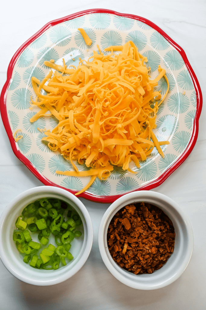 Two white bowls, one filled with chopped green onions and the other filled with bacon bits with a saucer full of shredded cheddar cheese. 