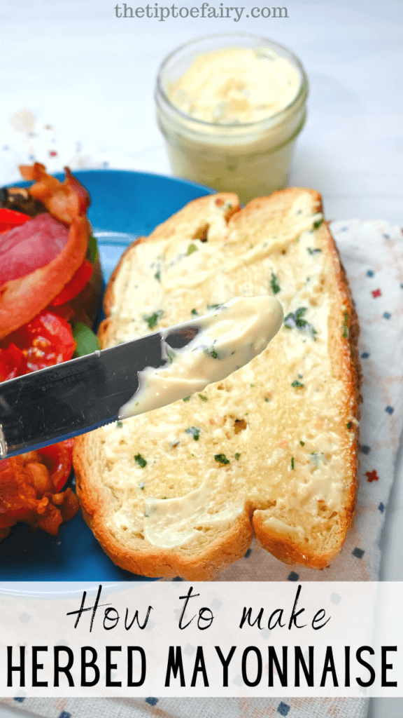 Close up of a knife with herbed mayonnaise on it and a sandwich behind it laid flat with the mayonnaise on one piece of bread. 