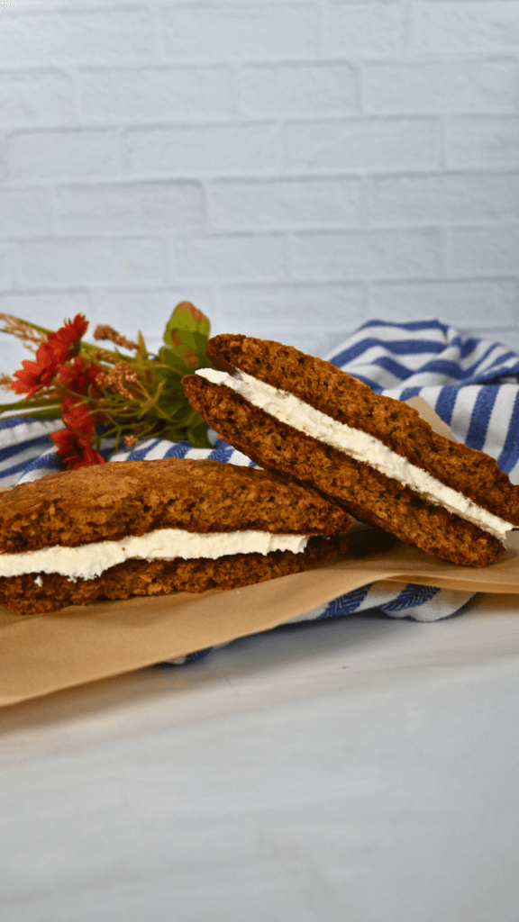 A giant oatmeal cream pie cut in half on a piece of parchment paper with flowers behind