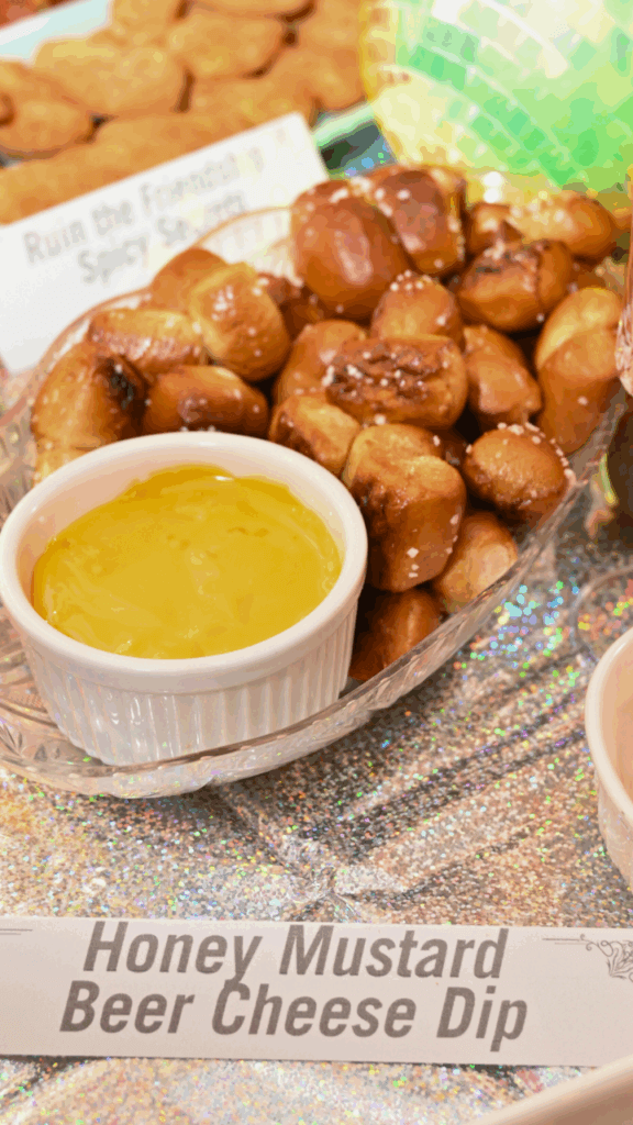 A close up of a small white bowl of honey mustard beer cheese dip surrounded by soft pretzel nuggets in a clear dish over an iridescent table cloth