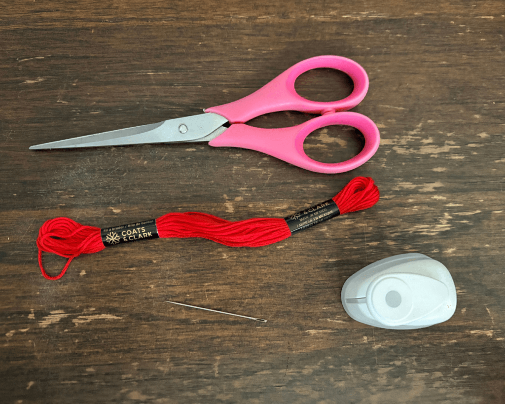 Supplies to make a bookmark tassel - a hole punch, red embroidery floss, a needle, and pink scissors. 