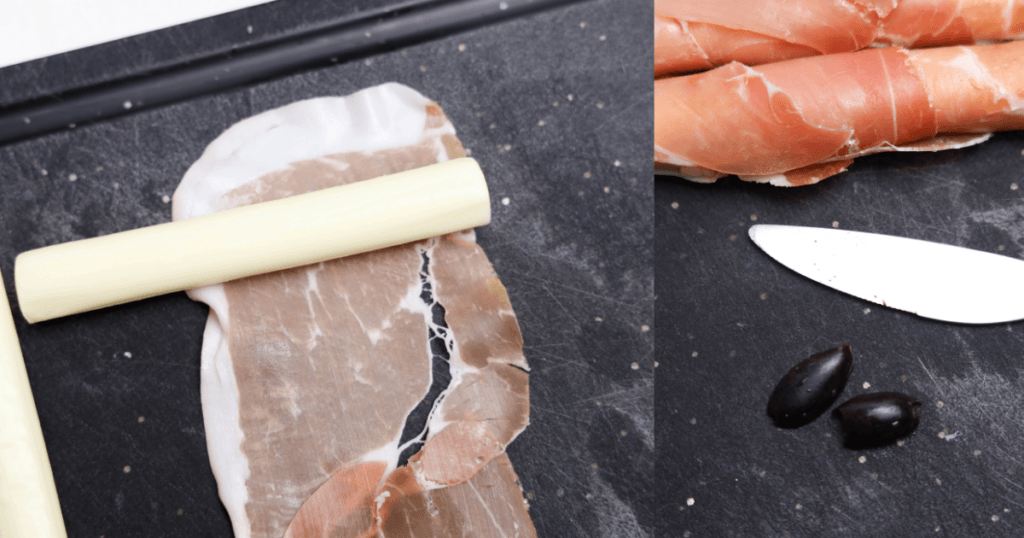Rolling up the string cheese with the prosciutto and cutting the olives into witch nails on a black cutting board. 